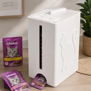 Op de afbeelding: Een witte plastic dispenser voor kattenvoerzakjes met een verticaal kijkvenster, een in reliëf gemaakte katsilhouet aan de zijkant en een deksel in de vorm van een kat. De dispenser bevat meerdere paarse Whiskas kattenvoerzakjes.