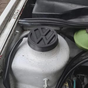 May include: A black circular cap featuring the Volkswagen logo embossed on the top surface. The cap is installed on a white cylindrical automotive component within an engine bay, surrounded by black wiring and a green reservoir container.