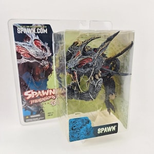 May include: A Spawn Mutations Series 23 action figure encased in clear plastic packaging. The creature features a dark, textured body with red accents, sharp spikes, and a silver chain. The packaging displays the text SPAWN.COM and SPAWN.