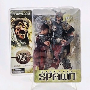Puede incluir: Una figura de acción de Spawn The Bloodaxe de la serie The Viking Age en su embalaje de plástico original. La figura presenta un guerrero esquelético con casco con cuernos, detalles de calaveras, cadenas y una capa roja. El embalaje incluye el texto SPAWN.COM, THE VIKING AGE y DARK AGES SPAWN.