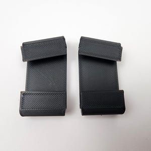 May include: Two black, rectangular, 3D-printed plastic clips with a textured, diagonal-line finish. Each clip features a flat central panel with a folded top hook and a bottom ledge, designed for mounting or securing items in a vertical position.