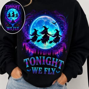 Peut inclure: Un sweat-shirt noir orné d'un motif représentant trois sorcières volant sur des balais devant une grande pleine lune bleue. Le design inclut des nuages violets et bleus, une forêt sombre et le texte TONIGHT WE FLY en lettres bleues.