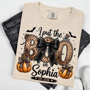 Peut inclure: Un t-shirt beige avec un motif portant le texte I put the BOO in Sophia EST. 2026. Le design comprend de grandes lettres à motif damier marron, un nœud noir, deux citrouilles orange avec des nœuds léopard, des chauves-souris et des toiles.