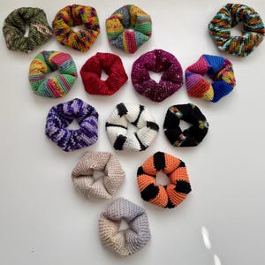 Op de afbeelding: Een verzameling handgemaakte scrunchies in verschillende kleuren en patronen. De scrunchies zijn gemaakt van gebreide stof en bevatten kleuren als rood, paars, oranje, zwart en wit. Sommige scrunchies hebben gestreepte ontwerpen, andere zijn effen.