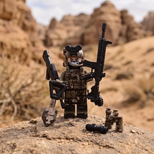 May include: A plastic toy soldier minifigure wearing a camouflage uniform, tactical vest, and a helmet with night vision goggles. The figure holds a black rifle and a shovel, with a pair of camouflage binoculars and a small black cylinder nearby.