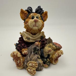 May include: A textured resin figurine of an orange cat wearing a dark purple outfit with a cream-colored collar and a black bow on its head. The cat is sitting and holding a gray knitted bag, with a smaller sleeping cat figure resting beside it.