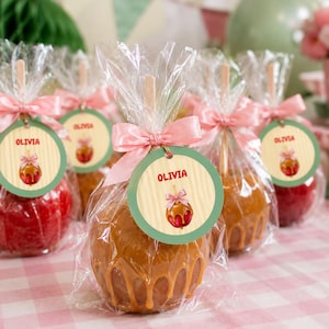 Puede incluir: Varias manzanas de caramelo en palitos de madera, envueltas individualmente en plástico transparente con un lazo de satén rosa. Cada una tiene una etiqueta redonda con fondo de rayas amarillo claro, el nombre OLIVIA y una ilustración.