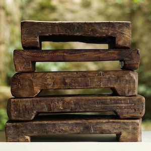 May include: A stack of five rectangular dark brown wooden risers, each featuring a thick, weathered surface with visible grain and short, sturdy feet at the corners, creating a tiered structure with a rough, natural texture and deep brown tones.