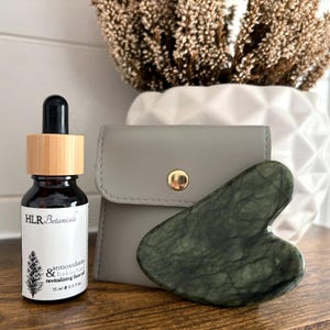 May include: A dark amber glass bottle of HLR Botanicals antioxidants and bakuchiol revitalizing face oil with a bamboo dropper cap, a grey faux leather pouch with a gold snap, and a heart-shaped dark green marbled stone gua sha tool.