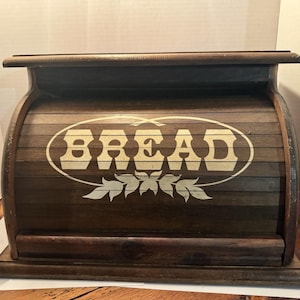 May include: A dark brown wooden bread box with a roll-top door. The front features the word BREAD printed in a cream-colored serif font inside an oval border, with a decorative leaf graphic positioned directly underneath the text.