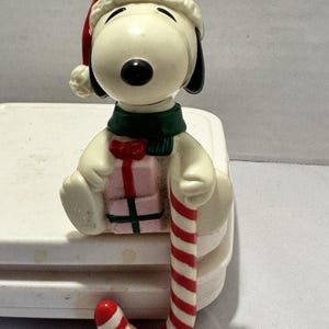 May include: A cream-colored dog figurine wearing a red Santa hat and a green scarf. The dog is sitting and holding a small pink gift box with a red bow and a green ribbon. A red and white striped candy cane hangs down from the dog's hand.