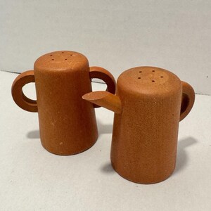 May include: Two terracotta-colored salt and pepper shakers with a matte finish. One shaker features a small spout and a single handle, while the other has two handles. Both shakers have a cylindrical body and a flat top with small holes for dispensing.