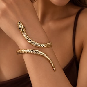 Peut inclure: Un bracelet serpent doré enroulé présentant un motif texturé en forme d'écailles sur toute sa longueur. La tête du serpent est détaillée avec de petits yeux sombres incrustés, et le corps s'enroule autour du bras dans un design en spirale.