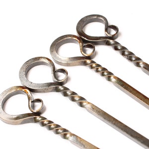 May include: Four metal skewers with twisted handles. The handles are shaped like a heart with a small loop at the top. The skewers are made of a dark metal with a rustic finish.