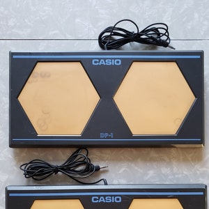 May include: Two black Casio DP-1 drum pads with two hexagonal orange pads each. The pads have blue trim and the word CASIO is printed in blue. Each pad has a black cable attached.