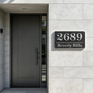 Könnte beinhalten: Ein rechteckiges Hausnummernschild mit strukturiertem schwarzem Hintergrund und silbernem Rand. Das Schild zeigt die silbernen Ziffern 2689 über dem Text Beverly Hills in einer Serifenschrift, montiert an einer hellgrauen Fliesenwand neben einer dunklen Tür.
