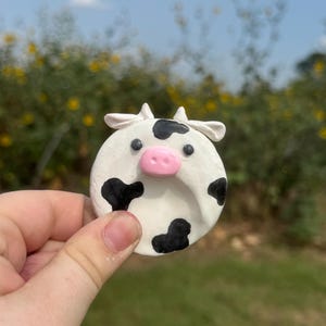 May include: A circular white clay ornament featuring a black cow-spotted pattern, two small white horns, and a raised pink snout. The object has two small black dots for eyes and a smooth, rounded surface, presenting a stylized animal face design.