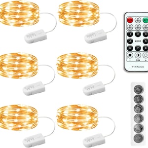 May include: Six coils of warm white LED fairy lights on thin copper wire, each connected to a small white battery pack. Includes a white rectangular remote control with numbered mode buttons and timer settings, plus a strip of six round coin batteries.