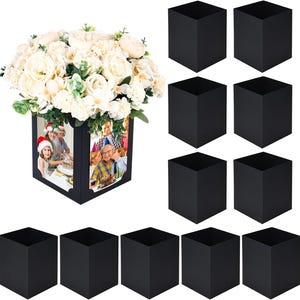 Puede incluir: Un conjunto de once jarrones cuadrados negros, uno de los cuales está lleno de un gran ramo de flores artificiales de color crema y presenta dos inserciones fotográficas rectangulares en sus lados que muestran personas en una fiesta navideña y un cumpleaños.