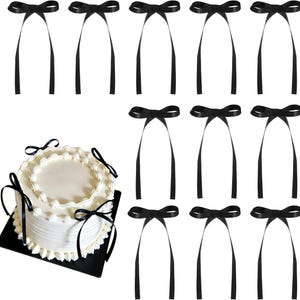 May include: A white round cake with white frosting and decorative piping, adorned with three black ribbon bows. Beside the cake are eleven additional black ribbon bows, each featuring two long, thin, trailing ribbon tails hanging downwards.