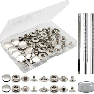 May include: A set of silver-toned metal snap fasteners in a clear plastic storage container, accompanied by three steel setting tools and a cylindrical base. The fasteners consist of smooth caps, socket components, and stud pieces for fabric assembly.