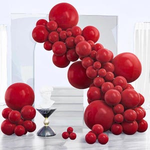 Puede incluir: Un gran arco de globos en cascada compuesto por numerosos globos rojos brillantes de varios tamaños. El arreglo presenta un grupo denso de esferas que crean una forma curva y orgánica, acompañado por un grupo más pequeño y separado de globos rojos.