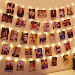 Puede incluir: Una cadena de luces LED de color blanco cálido con numerosos clips pequeños que sostienen varios pósteres rectangulares de estilo vintage, fotografías en blanco y negro e impresiones coloridas de viajes en una pared.