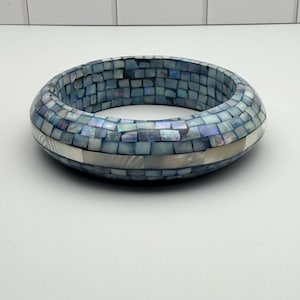 May include: A circular bangle bracelet featuring a mosaic pattern of iridescent blue and white shell tiles. The surface is composed of small, square-shaped pieces arranged in rows, with a central horizontal band of elongated, smooth white shell segments.