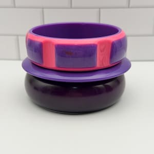 May include: A stack of three bangle bracelets. The top bangle is purple with a bright pink horizontal stripe. The middle bangle is a thin, flat purple ring. The bottom bangle is a wide, solid dark purple band with a smooth, glossy finish.