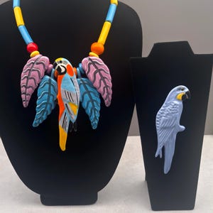 May include: A necklace featuring a central parrot pendant surrounded by pink and blue leaf-shaped charms, suspended from a strand of yellow, blue, and red cylindrical beads, alongside a separate light blue parrot brooch on a display stand.