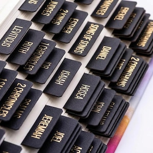 May include: A set of black adhesive index tabs for a Bible, featuring gold foil lettering for various book names including Leviticus, Ruth, 1 Chronicles, Job, Isaiah, Hosea, Jeremiah, Joel, Daniel, 2 Corinthians, and Song of Songs.