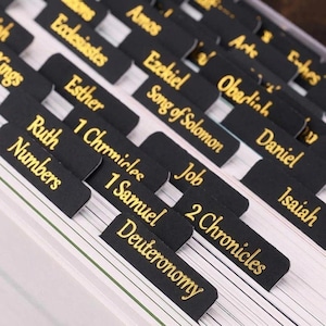 May include: A set of black adhesive bible index tabs with gold lettering. The tabs feature names of various books of the Bible including Deuteronomy, 1 Samuel, 2 Chronicles, Numbers, Ruth, 1 Chronicles, Job, Esther, Song of Solomon, Ezekiel, and Isaiah.