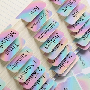 May include: A collection of adhesive Bible index tabs featuring a holographic, iridescent gradient finish in shades of pink, blue, and purple. Each tab is printed with black text identifying specific books of the Bible.