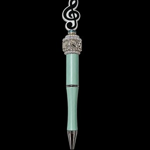 May include: A light mint green ballpoint pen featuring a silver-toned metal tip and a decorative topper. The topper includes a white and black treble clef symbol, a band of small white faux pearls, and clear faceted crystals set in silver.