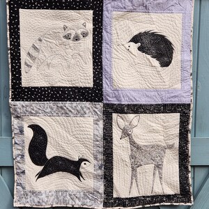 May include: A black and white quilt with four squares featuring different animals. The top left square has a raccoon, the top right square has a hedgehog, the bottom left square has a squirrel, and the bottom right square has a deer.
