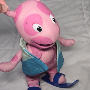 May include: A pink plush character with darker pink spots, wearing a light blue vest and dark blue plastic skis on its feet. The character has large white eyes with black pupils, two antennae on its head, and a soft, stuffed fabric body construction.