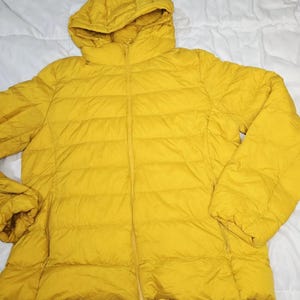 May include: A bright yellow puffer jacket featuring a quilted design, a full-length front zip, and an attached hood. The jacket has long sleeves with elasticated cuffs and two side pockets with zip closures, providing a consistent appearance.