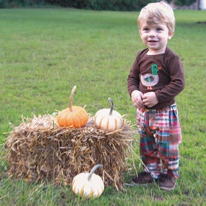 May include: A young boy wearing a brown long-sleeved shirt with a green duck applique and plaid pants stands in a field with a bale of hay and pumpkins.