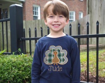 May include: A navy blue long-sleeved shirt featuring a turkey design with a plaid pattern and the name "Jude" embroidered in gold. The shirt is paired with brown corduroy pants. The boy is smiling.