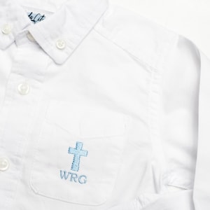 May include: White button-down shirt with a light blue embroidered cross on the pocket. The shirt has a button-down collar and long sleeves. The initials "WRG" are embroidered below the cross.