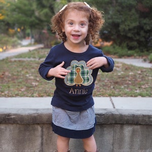 May include: A young girl wearing a blue long-sleeved shirt with a turkey applique and the name "Annie" embroidered on it. She is also wearing a plaid skirt and brown cowboy boots.