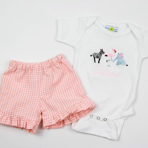 K&ouml;nnte beinhalten: Eine rosa-wei&szlig;e Gingham-R&uuml;schenhose und ein wei&szlig;er Strampler mit einem gestickten Design aus Zebra, Flamingo und Elefant. Der Strampler ist mit dem Text "Sullivan" bestickt.