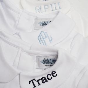 May include: Close-up of three white baby rompers with Peter Pan collars. Each romper has embroidered text: "RLP III", "RPL", and "Trace". The rompers have tags that read "Soda City Sewing".