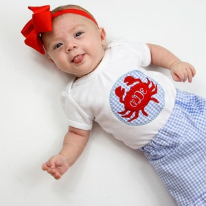 Op de afbeelding: Een baby draagt een wit rompertje met een rode krab-applicatie en het monogram "A.D.E." Het rompertje wordt gecombineerd met blauw-wit geruite shorts. De baby draagt een rood strikband.