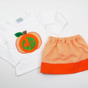 Op de afbeelding: Een wit longsleeve shirt met een groene en oranje pompoen applique met een monogram. Het shirt wordt gecombineerd met een wit en oranje gingham rok met een oranje band.
