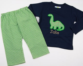 Puede incluir: Una camisa de manga larga azul marino con una aplicación de dinosaurio de vichy verde y el nombre "Jake" bordado en marrón. La camisa se combina con pantalones de vichy verde.