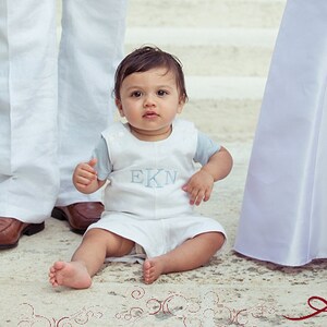 May include: A young boy in a white and blue outfit sits on a white tiled floor. He is wearing a white shirt with blue trim and the letters "EKN" embroidered on the front. The boy is looking at the camera with a serious expression. Two adults, a man and a woman, are standing behind the boy, holding hands. The man is wearing a white suit and the woman is wearing a white dress.
