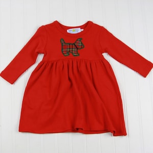 Könnte beinhalten: Rotes Langarmkleid mit geraffter Taille. Das Kleid hat einen Applikation eines Hundes mit Karomuster auf der Brust. Das Kleid ist aus weichem Material und für Kinder geeignet.