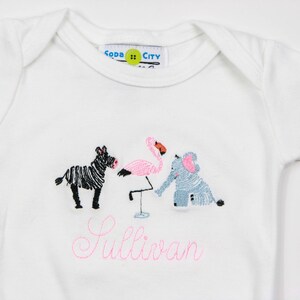 K&ouml;nnte beinhalten: Wei&szlig;er Baby-Strampler mit rosa Stickerei eines Zebras, Flamingos und Elefanten. Der Strampler hat den Text "Sullivan" in rosa gestickt.