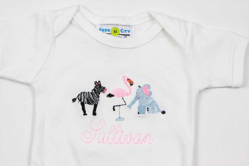 K&ouml;nnte beinhalten: Wei&szlig;er Baby-Strampler mit rosa Stickerei eines Zebras, Flamingos und Elefanten. Der Strampler hat den Text "Sullivan" in rosa gestickt.